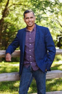 Simon Mittal in red shirt blue blazer and blue jeans, leaning against a fence