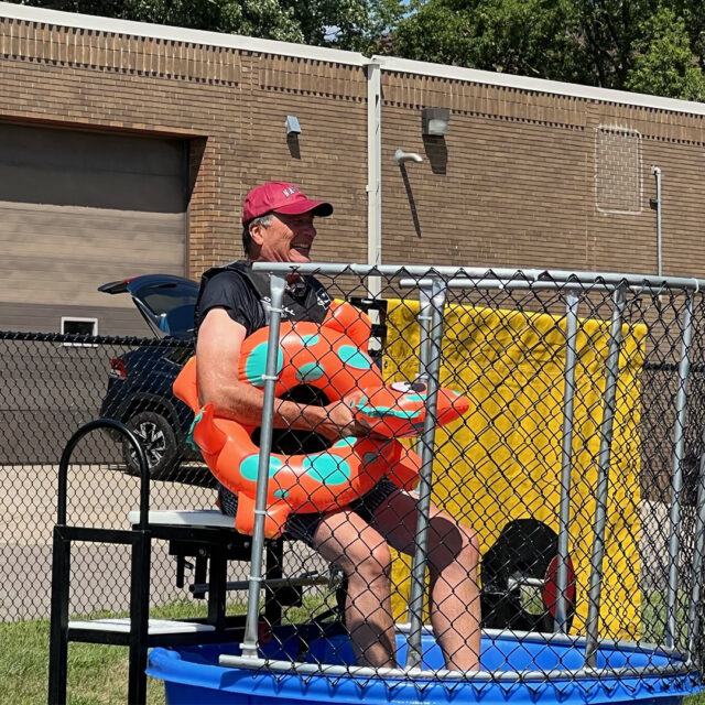 Cassia President and CEO Bob Dahl smiles while sitting in a dunk tank, wearing a life jacket and holding an orange dinosaur floatie during a State Fair-inspired picnic event.