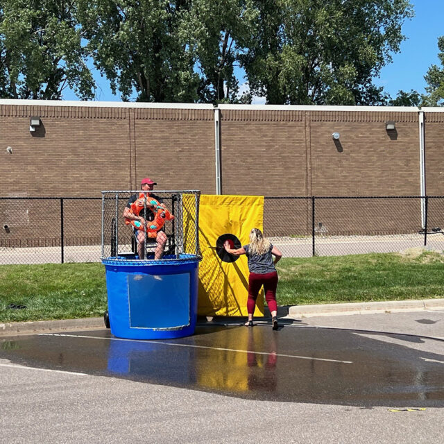 Cassia President and CEO Bob Dahl sits in a dunk tank, wearing a life jacket and holding an orange dinosaur floatie, as a colleague prepares to press the target during a State Fair-inspired picnic event.