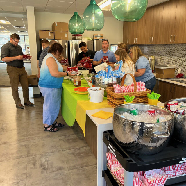 Cassia employees line up to prepare their own walking tacos at a food station during a State Fair-inspired picnic, with bowls of snacks, drinks, and sweet treats available on the table.