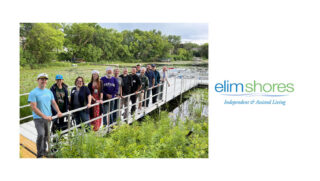 A group of smiling residents and staff from Elim Shores, an independent and assisted living community, stands on a dock next to a serene waterway surrounded by lush greenery. The group, dressed in casual attire, celebrates the new outdoor amenities, including a dock and pontoon boat, made possible by generous donations.
