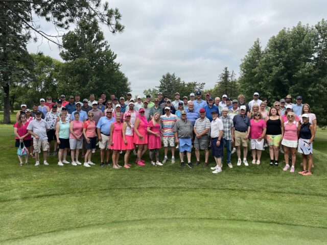 A large group of people gathered on a golf course, posing for a group photo at a golf tournament. They are dressed in a variety of golf attire, with many wearing shades of pink and casual outfits. The group includes a diverse mix of ages, all smiling and standing closely together. The lush green surroundings, including trees and a well-manicured lawn, provide a picturesque backdrop. The scene captures a cheerful and friendly atmosphere at the tournament.