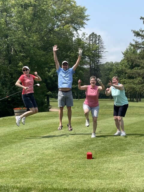 Four people jumping in the air on a golf course, clearly enjoying a playful moment. They are dressed in casual golf attire, with two women on the left and right, and two men in the middle. The scene captures a joyful, light-hearted vibe.