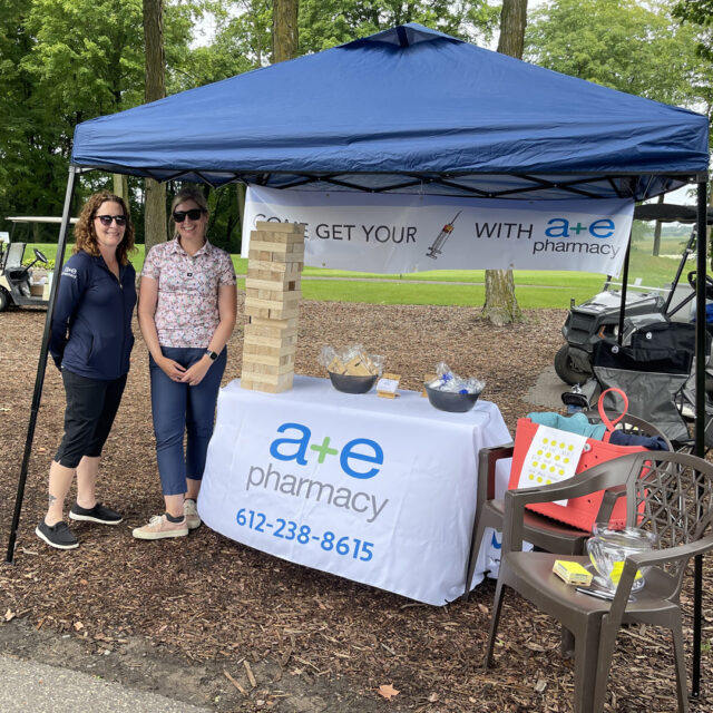 Two representatives from a+e Pharmacy stand under a tent at the 2024 Cassia Golf Tournament. They are next to a table with promotional items and a large Jenga game, with the company’s logo displayed on a banner and tablecloth. Golf carts and trees are visible in the background.