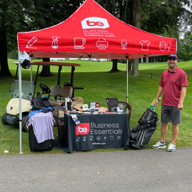A representative from Business Essentials stands next to a branded tent at the 2024 Cassia Golf Tournament. The tent displays the company’s logo and promotional items are arranged on a table underneath. A golf cart and golf bags are visible in the background on the green course.