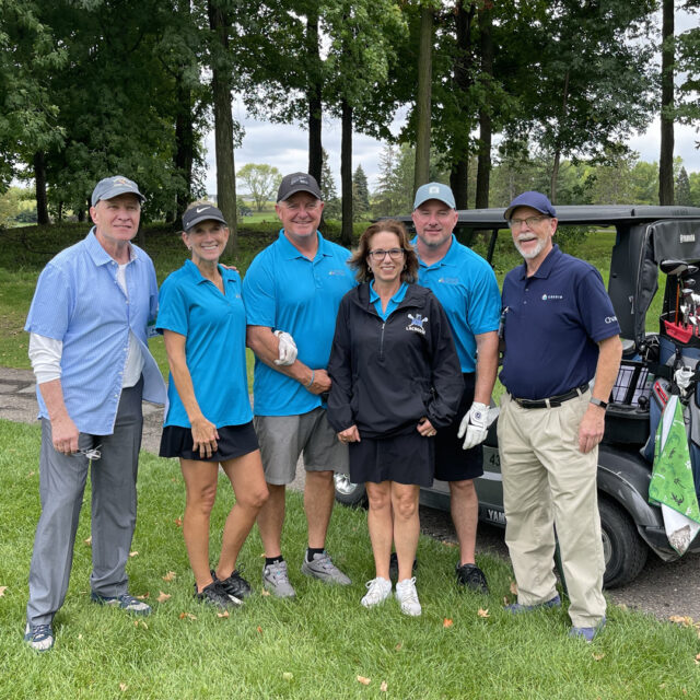 A group of six participants from the 2024 Cassia Golf Tournament pose together in front of a golf cart. They are wearing matching blue shirts, smiling, and standing on the green with trees in the background. The atmosphere is friendly and relaxed as they enjoy the tournament.