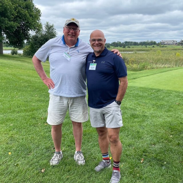 Two men smile and pose together on the green at the 2024 Cassia Golf Tournament. They are wearing casual golf attire and name tags, standing on a grassy area with a scenic view of greenery and a cloudy sky in the background.