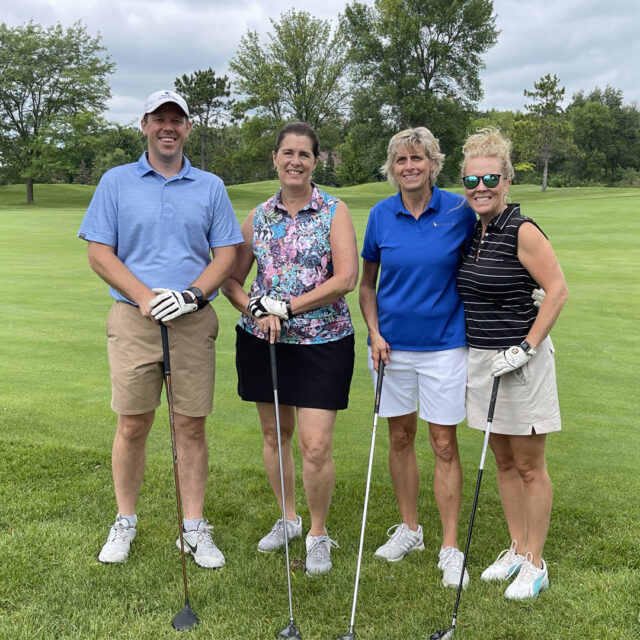 A group of four golfers pose together on the green during the 2024 Cassia Golf Tournament. They are smiling and holding their clubs, dressed in casual golf attire, with trees and a bright green course in the background.