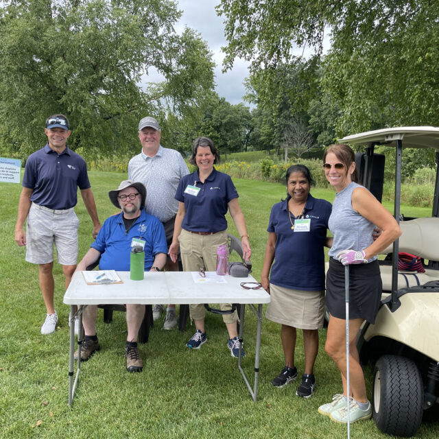 A group of six individuals pose together outdoors during the 2024 Cassia Golf Tournament. They are standing near a golf cart on the green, smiling and enjoying the event. One person is seated at a table with golf supplies, while others stand nearby, ready for the tournament.