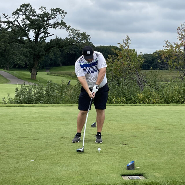 A golfer prepares to swing his club at the 2024 Cassia Golf Tournament. He is standing on the tee, focused on hitting the ball, with a green course and trees in the background under a cloudy sky.