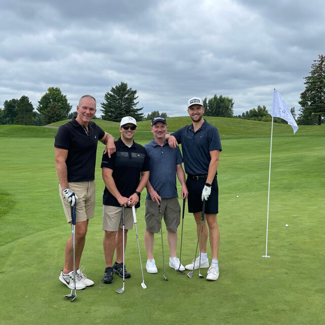 A group of four golfers pose together on the green during the 2024 Cassia Golf Tournament. They are smiling and holding their clubs, standing near a flag on the course. The cloudy sky and lush surroundings create a scenic backdrop as they enjoy the event.