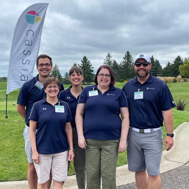 The Philanthropy team at Cassia poses together outdoors at the 2024 Cassia Golf Tournament. The group of five is smiling, wearing matching navy blue shirts with name tags, standing near a Cassia flag on a green golf course. The team is enjoying the event under a cloudy sky.
