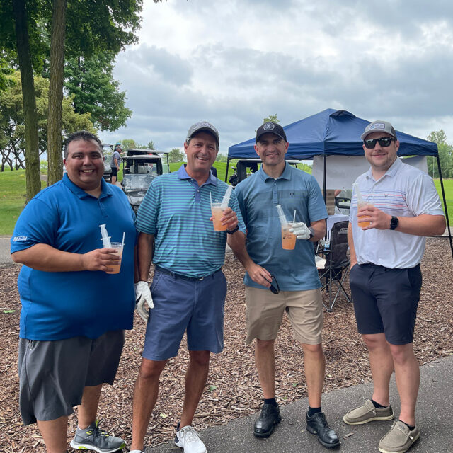Four participants at the 2024 Cassia Golf Tournament smile and hold drinks while standing outdoors near a tent and golf carts. They are casually dressed in golf attire, enjoying refreshments together during a break in the event.