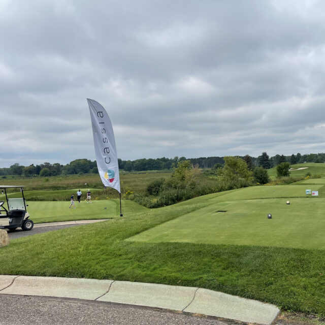 A scenic view of the golf course during the 2024 Cassia Golf Tournament, featuring a Cassia-branded flag near the tee box. A golf cart is parked nearby, and two golfers are seen in the distance preparing for their shots under a cloudy sky, with greenery and rolling hills in the background.