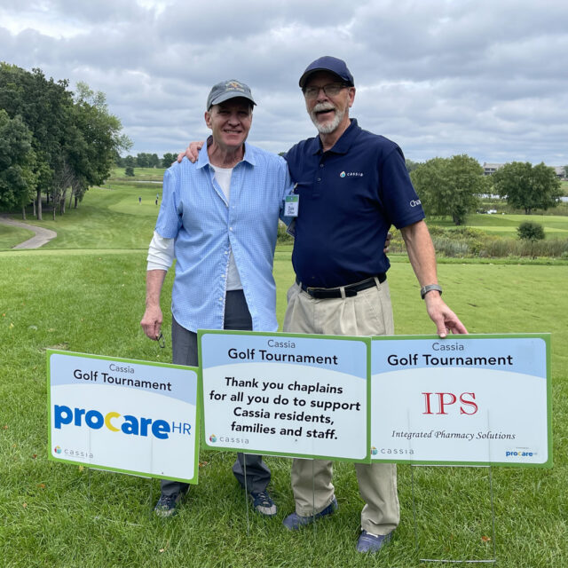 Two men at the 2024 Cassia Golf Tournament pose together on the golf course, standing behind signs thanking event sponsors. The signs feature messages of appreciation for chaplains and sponsors like ProCare HR and IPS. The green golf course and cloudy sky create a peaceful backdrop.