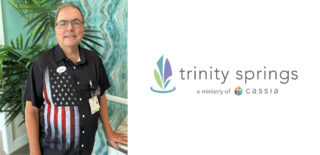 Rev. Chuck Tabor, Chaplain at Trinity Springs, stands smiling in a patriotic shirt, ready to share his compassionate spiritual care and support with the community.