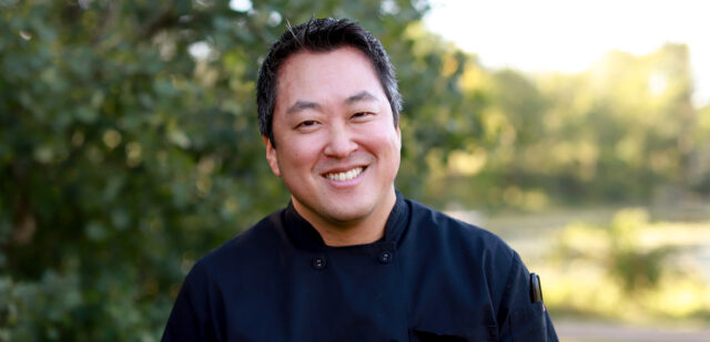 A portrait of Chef Ryan Schmitt, Director of Culinary Services at Elim Shores, smiling outdoors in natural light. He is wearing a black chef's jacket, standing against a blurred background of green trees and sunlight.
