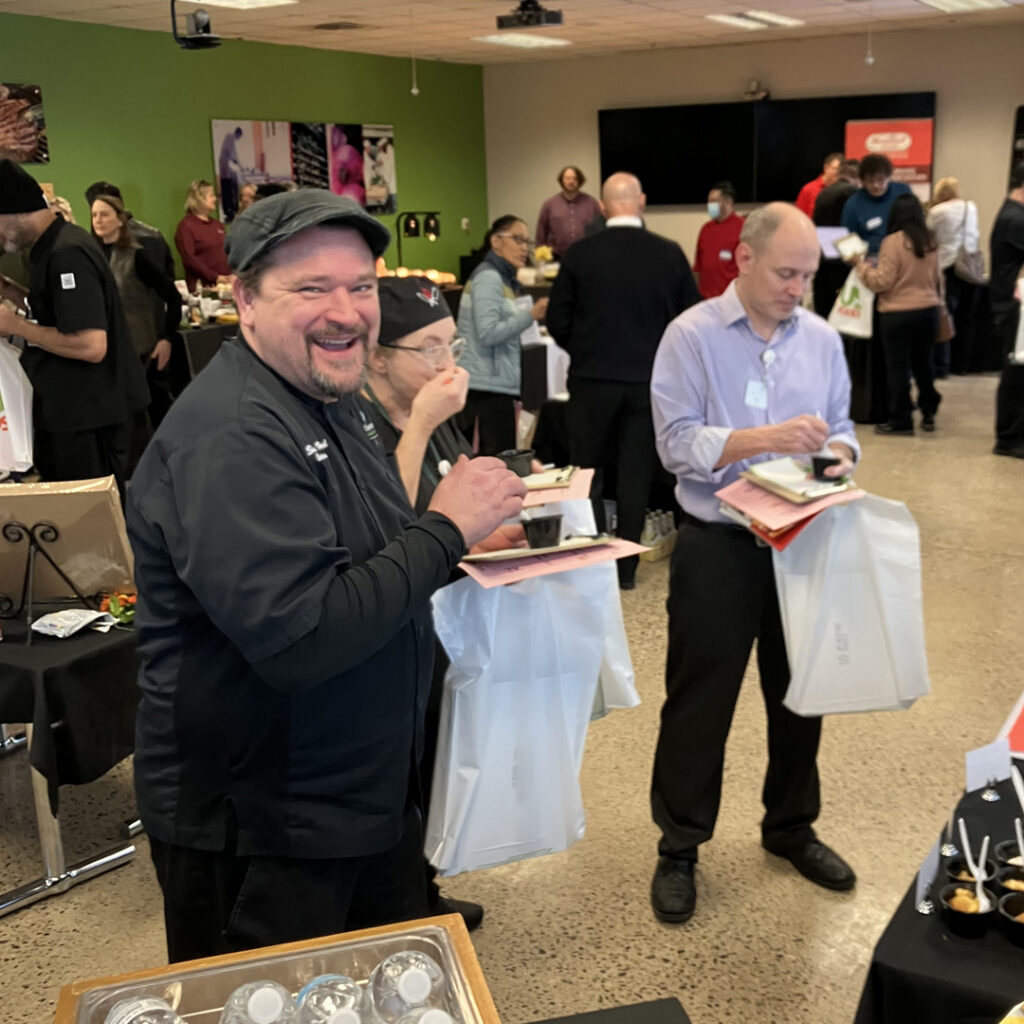 A smiling culinary team member at the Cassia food show enjoys tasting samples while taking notes, surrounded by colleagues and food vendors in a collaborative atmosphere.