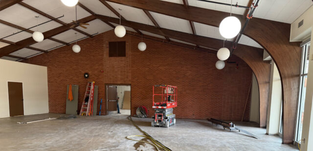 A spacious chapel under renovation with a brick wall, exposed ceiling beams, and construction equipment scattered throughout.