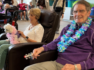 Memory Care residents at Trinity Springs enjoy live entertainment while wearing leis and relaxing in recliners, joined by a small therapy dog.