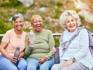Three older women sitting outdoors in warm weather, smiling and holding water bottles, representing the wellness, connection and comfort of a summer move to senior living in Cedar Rapids, Iowa.