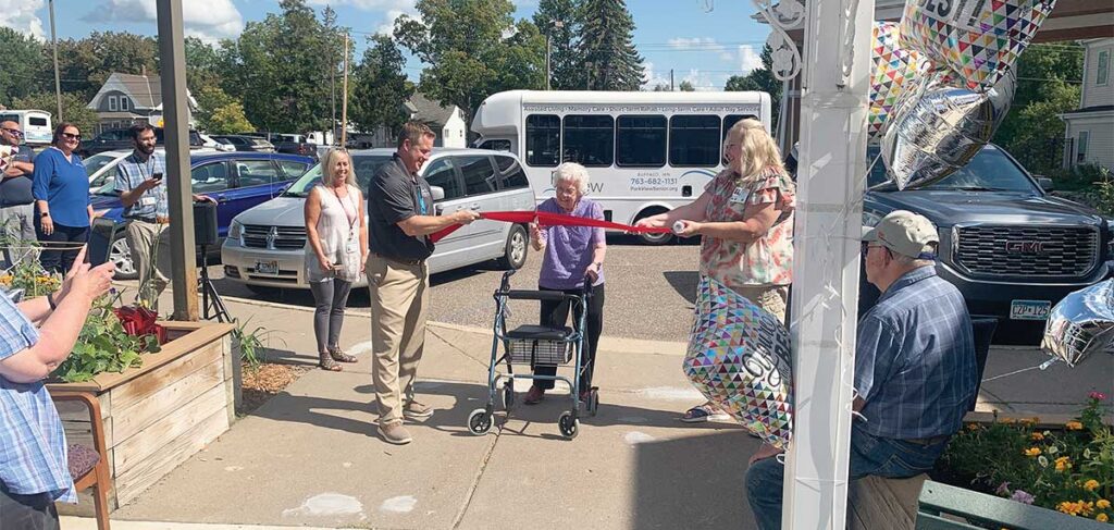 Resident cuts a ribbon during the celebration of a new donor-funded bus at Lake Ridge and Park View Assisted Living, surrounded by community members and balloons.