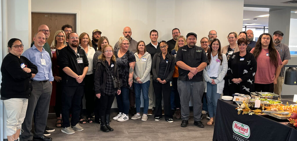 Cassia senior living culinary team gathered for a culinary training event, featuring chefs and dining staff standing together with a Hormel Foods foodservice display, highlighting teamwork and professional dining excellence.