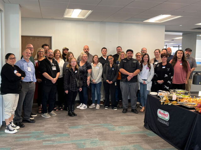 Cassia senior living culinary team gathered for a culinary training event, featuring chefs and dining staff standing together with a Hormel Foods foodservice display, highlighting teamwork and professional dining excellence.