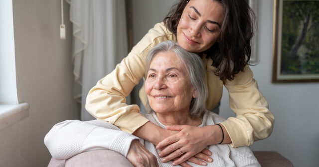 A person standing behind another, gently placing their arms around the seated individual