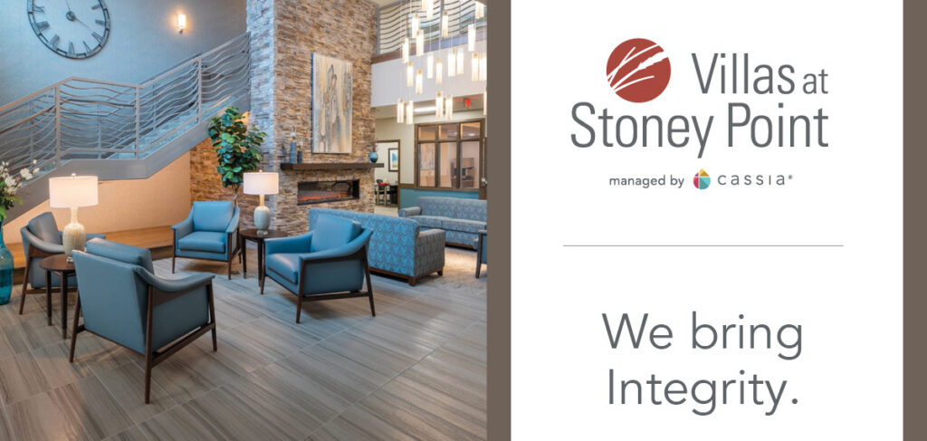 Lobby seating area at Villas at Stoney Point with chairs, fireplace, staircase, and community branding panel