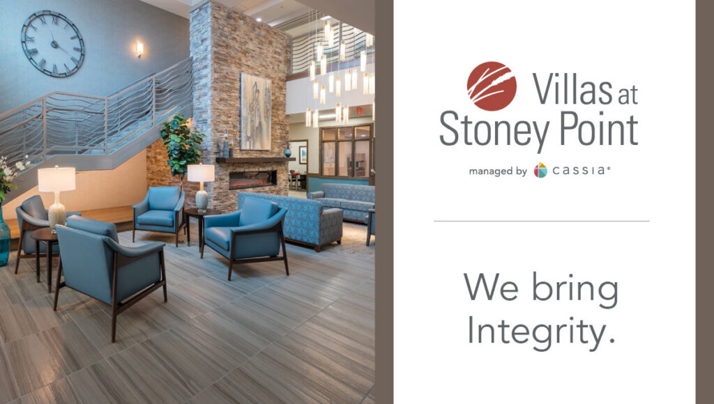 Lobby seating area at Villas at Stoney Point with chairs, fireplace, staircase, and community branding panel