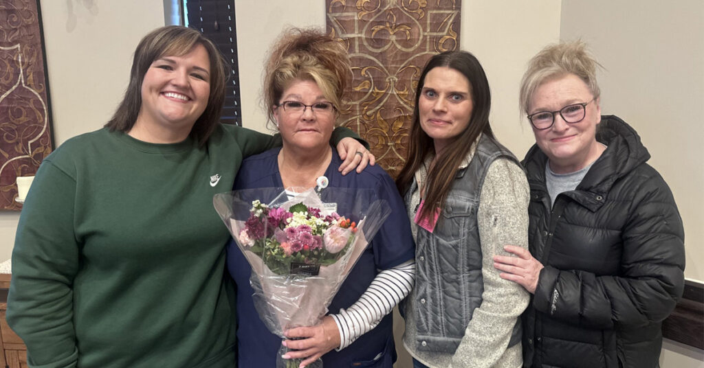 Group of four adults indoors, with the center person holding flowers while being recognized as Caregiver of the Year.