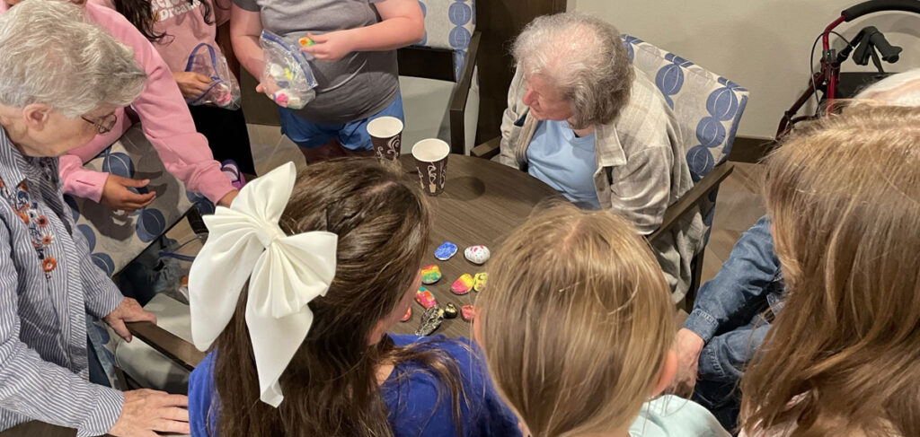 Residents sit around a table with a group of students as they look at and share colorful hand‑painted ‘happy rocks’ during an intergenerational activity