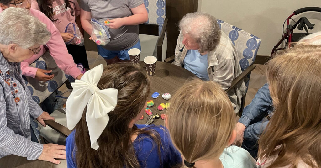 Residents sit around a table with a group of students as they look at and share colorful hand‑painted ‘happy rocks’ during an intergenerational activity