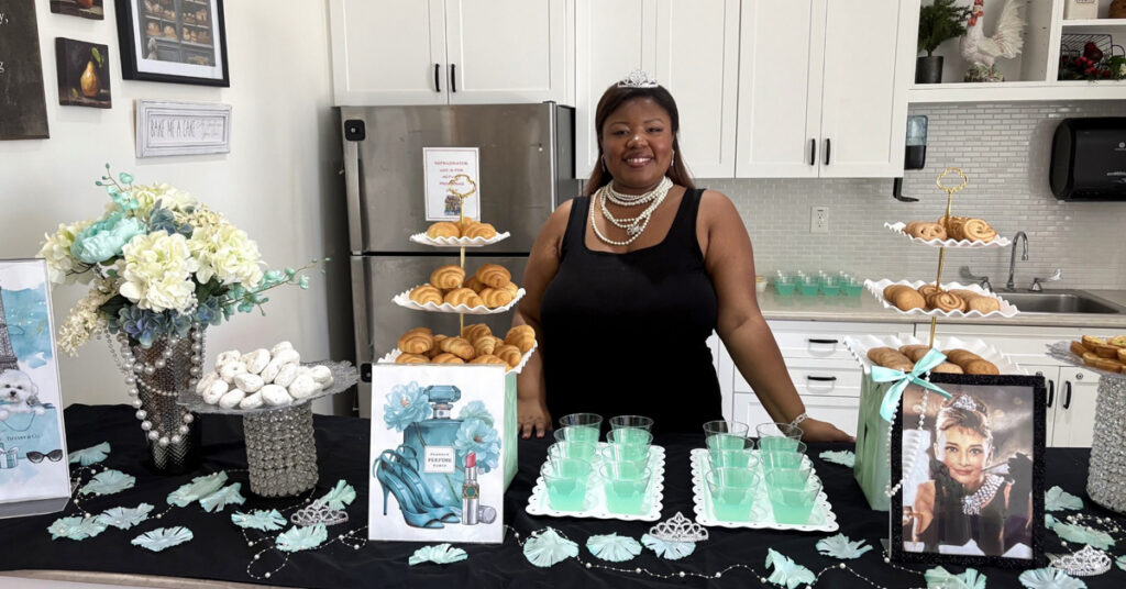 An employee standing behind a decorated refreshment table at a senior living event, featuring pastries, teal drinks, flowers, and themed decor with an Audrey Hepburn image in a bright kitchen setting.