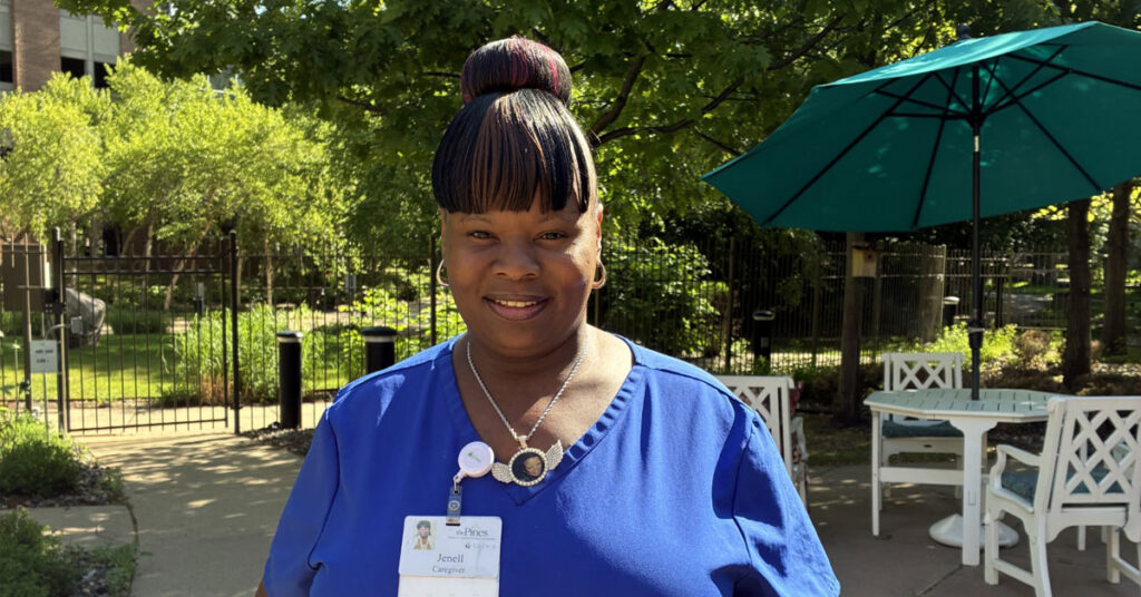 Caregiver at The Pines senior living community standing outdoors in a landscaped courtyard with patio seating and greenery, wearing blue scrubs and a name badge.
