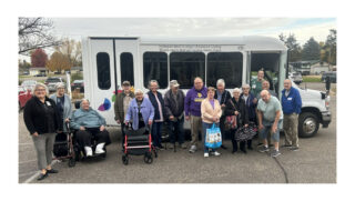 Group of senior living residents and staff standing together outdoors in front of a bus labeled for independent living, assisted living, and rehab services, with some using walkers and holding bags.