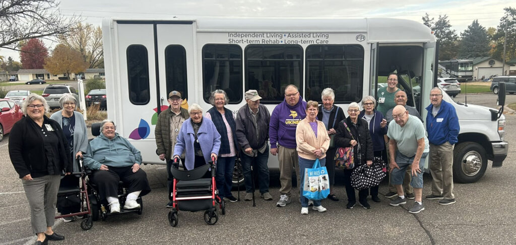 Group of senior living residents and staff standing together outdoors in front of a bus labeled for independent living, assisted living, and rehab services, with some using walkers and holding bags.