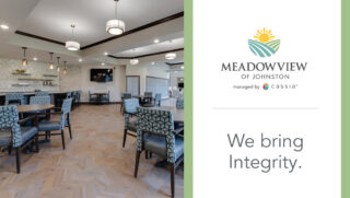 Interior common area at Meadowview of Johnston featuring dining tables, upholstered chairs, a bar seating area, and a television in a bright, modern senior living community. Branding on the image reads “Meadowview of Johnston” and “We bring Integrity.”