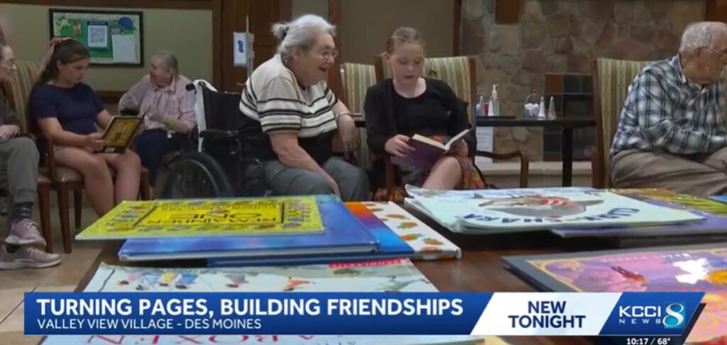 Elementary student reading a book with an assisted living resident at Valley View Village in Des Moines during an intergenerational literacy program, with other students and residents reading together in the background.