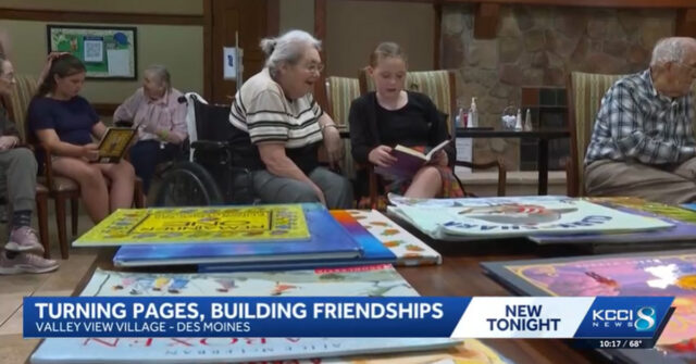 Elementary student reading a book with an assisted living resident at Valley View Village in Des Moines during an intergenerational literacy program, with other students and residents reading together in the background.