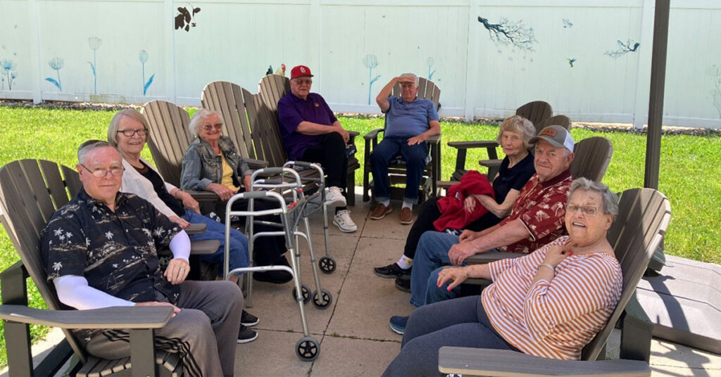 Residents gathered outdoors at a senior living community in Adirondack chairs, sitting in a circle on a patio area with green lawn and a white fence in the background.