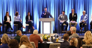 Panel discussion at a professional event with multiple speakers seated on stage and one speaker presenting at a podium, in front of an audience seated at round tables.