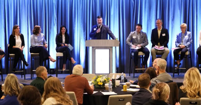 Panel discussion at a professional event with multiple speakers seated on stage and one speaker presenting at a podium, in front of an audience seated at round tables.