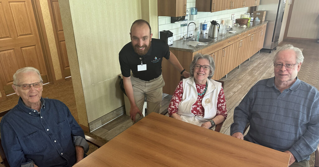 Chaplain at Cornerstone Assisted Living visiting with three residents seated around a table in a community common area with kitchen cabinetry and countertops in the background.