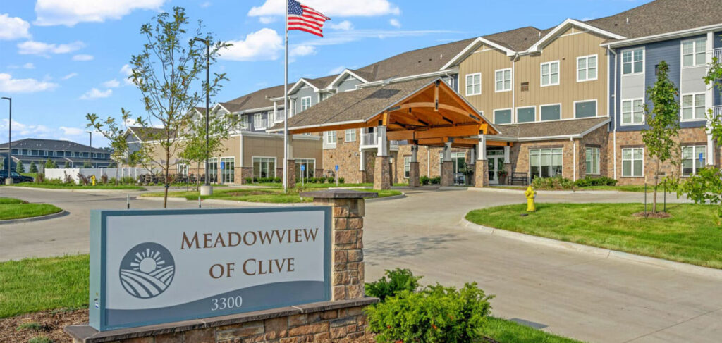 Exterior entrance of Meadowview of Clive senior living community featuring a covered entrance, landscaped grounds, American flag, and community sign displaying the Meadowview of Clive name.