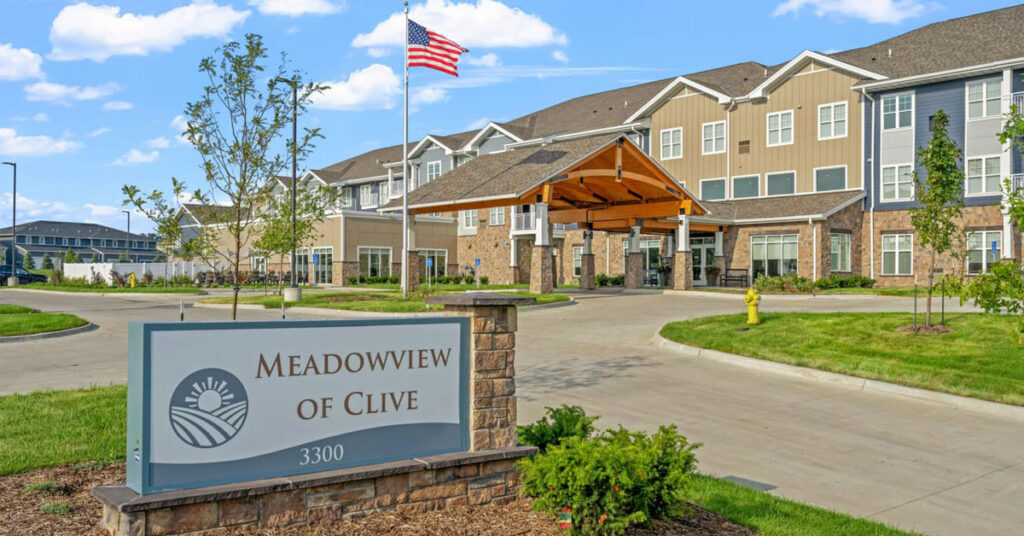 Exterior entrance of Meadowview of Clive senior living community featuring a covered entrance, landscaped grounds, American flag, and community sign displaying the Meadowview of Clive name.