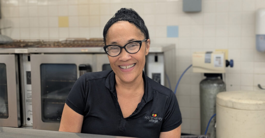 Team member at Apple Valley Village standing in a commercial kitchen area, wearing a black Apple Valley Village polo shirt, with ovens and food service equipment visible in the background.