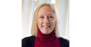 Portrait of a person wearing a red turtleneck sweater and dark cardigan, standing indoors in front of softly blurred windows.