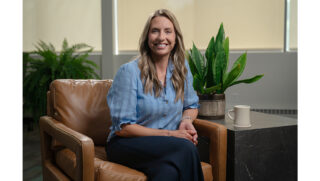 Person seated in a tan armchair in an office lounge with plants and a coffee mug nearby.